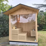 · Puerta semi cerrada con una pequeña ventanita para asegurar privacidad en uno de los laterales. · Cristal panorámico gigante de 2×2 metros, completamente continuo y sin cortes, que abre la vista del interior al jardín. · Tejado amplio estilo caseta, sobresaliente y protector, que refuerza la estética exterior y la funcionalidad frente a la intemperie. “Una sauna exterior a medida que combina privacidad, diseño panorámico y el máximo confort para disfrutar del bienestar en casa.”